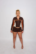 A woman stands against a light gray studio background wearing a matching dark brown, white, and orange athletic zip-up jacket and shorts set with 'Wynher' and number 8 graphics.