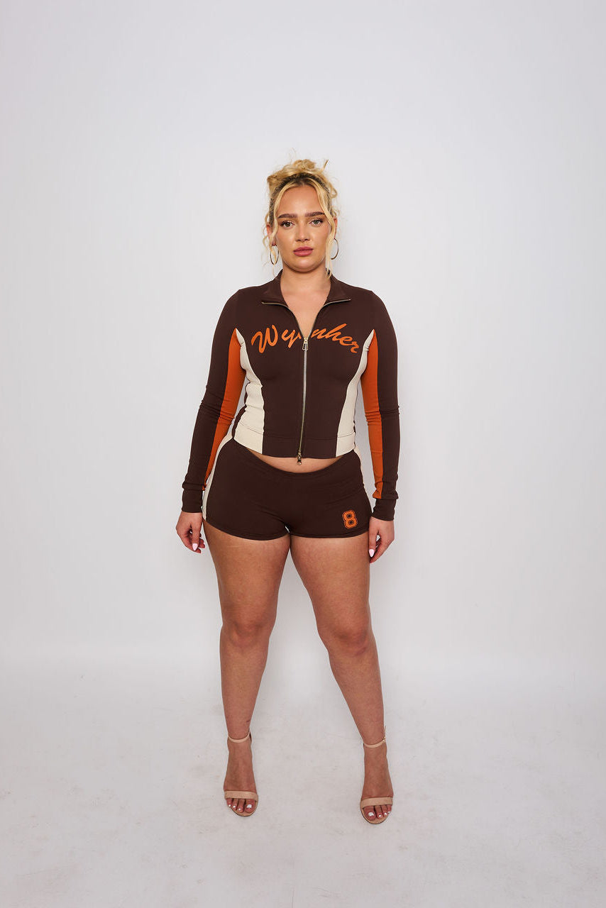 A woman stands against a light gray studio background wearing a matching dark brown, white, and orange athletic zip-up jacket and shorts set with 'Wynher' and number 8 graphics.