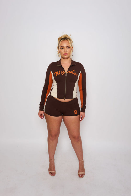 A woman stands against a light gray studio background wearing a matching dark brown, white, and orange athletic zip-up jacket and shorts set with 'Wynher' and number 8 graphics.