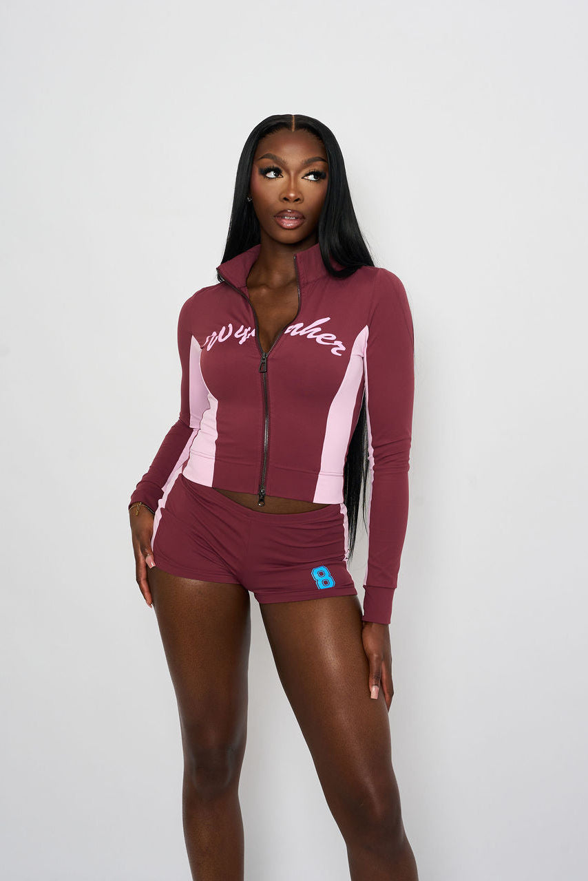 A woman modeling a matching burgundy and light pink athletic set consisting of a zip-up jacket and shorts, against a plain light grey background.