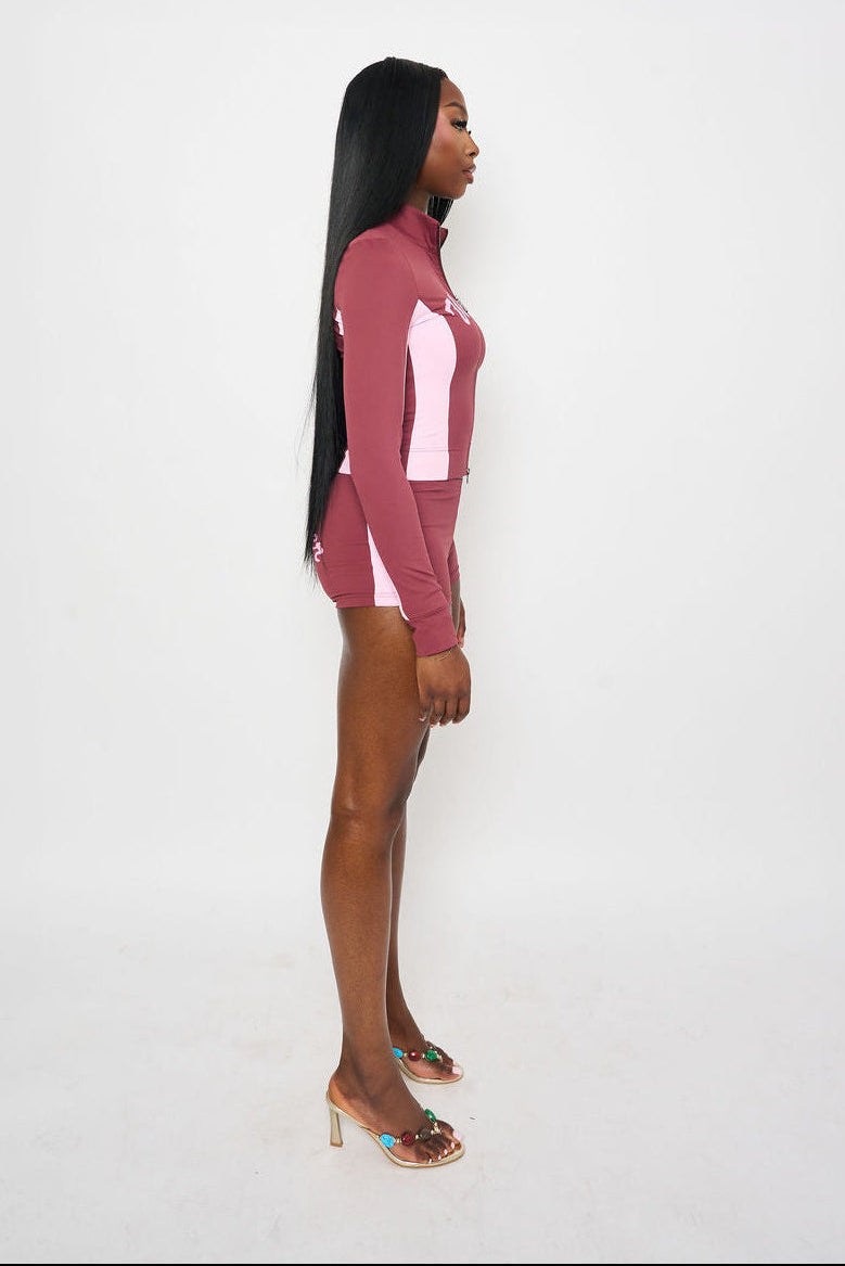 A woman stands in side profile wearing a long-sleeve athletic full-length jacket and matching shorts in burgundy and light pink, paired with clear high-heeled sandals, against a plain white studio background.