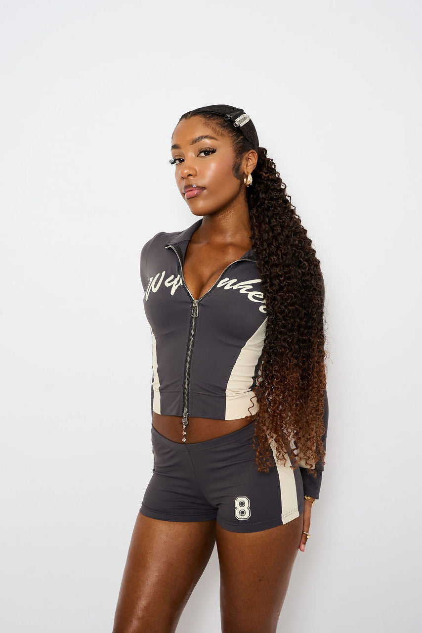 A woman modeling a charcoal and cream athletic two-piece set consisting of a cropped zip-up jacket and matching shorts, set against a plain white studio background.