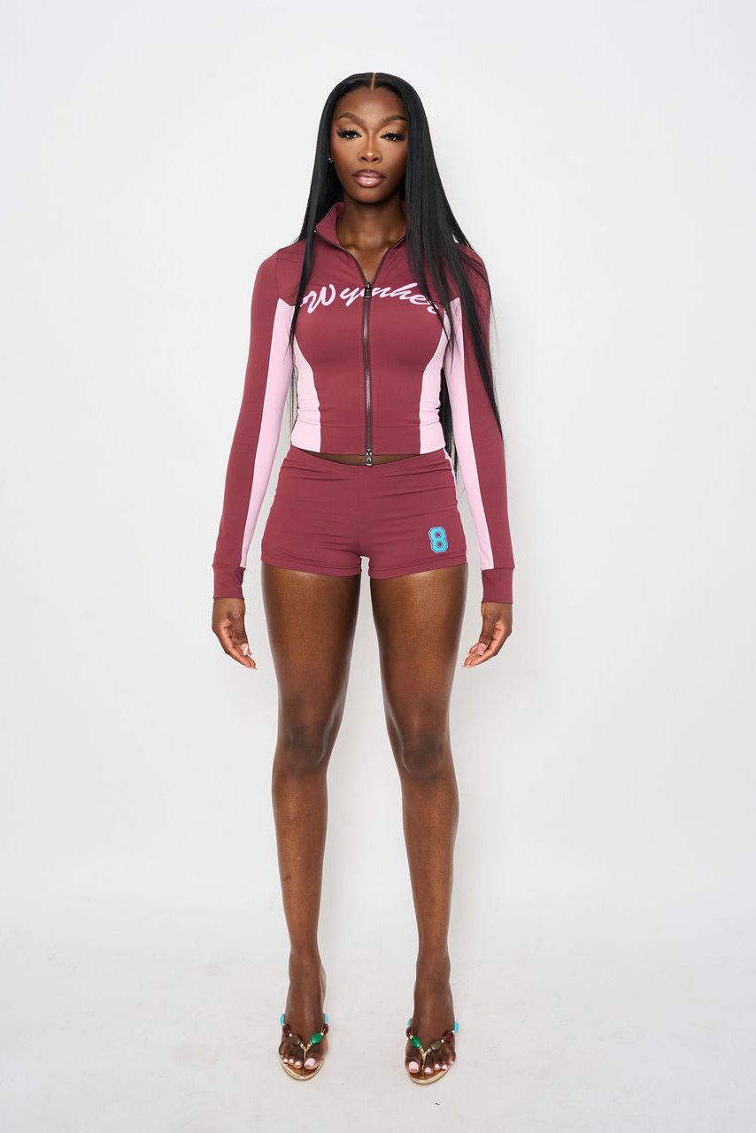 A woman modeling a matching two-piece athletic set consisting of a long-sleeve zip-up jacket and shorts in burgundy and light pink, with white script lettering 'WYNHER' and a small blue number 8 on the shorts, set against a plain light grey background.